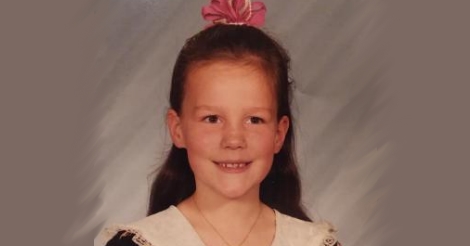 A school photo of Erica when she was about eight years old. She is smiling peacefully, with a hint of mild concern behind her eyes. She has a hair bow on top of her head, her long hair is pulled back, her little dress has some sort of laced shawl attached to it in that 1990s little girl fashion they used to have.