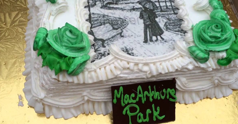 This is a real photo from somewhere of a pretty enough white cake with green roses on it. There is also a drawing of two people in a park on it. And in front of the cake, it is labeled "MacArthurs Park."