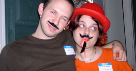 An old almost-blurry photo of a guy and a gal -- Kevin and Amandoll. They both have various amounts of fake facial hair on, and name tags. Amandoll has fake blood streaming from her mouth and a matching red hat.