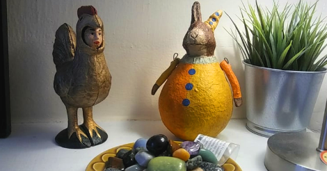 Photo of a cast iron chicken with a human face out of its mouth, standing with a rabbit sculpture that is shaped like an egg and dressed like a clown. In front of them is a big bowl of gemstones.